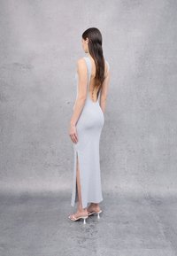 Vestido maxi gris ajustado con un profundo escote en la espalda, aberturas laterales y tela texturizada. Combinado con sandalias de tacón blancas, la modelo se encuentra frente a un fondo gris.