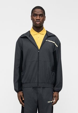 Homme portant une veste zippée noire à motif quadrillé et un pantalon assorti sur un polo jaune, debout devant un fond uni.