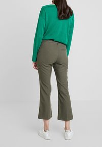 Žena oblečená v olivově zelených zkrácených kalhotách, zeleném dlouhém rukávu a bílých teniskách, stojící před jednoduchým světlým pozadím.