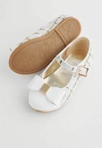 Weiße Mary Jane Schuhe mit einem glänzenden Bogen und goldenen Akzenten. Der Oberstoff ist aus einem strukturierten Gewebe und die Gummisohle ist gemustert und mit Schleifen versehen.