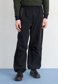 Černé, volné kalhoty vyrobené z lehkého materiálu s elastickým pasem a boční kapsou na zip. Nosené s černými botami.