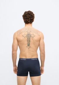 Boxers elásticos de algodão azul marinho com cós branco com logótipo, usados por um modelo masculino; tatuagem detalhada visível nas costas.