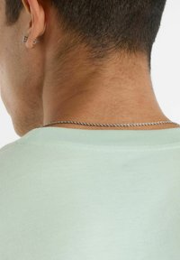 T-shirt di cotone verde chiaro vista da dietro con scollatura a costine, che mostra una texture liscia e una sottile catena argentata attorcigliata posata sul collo.