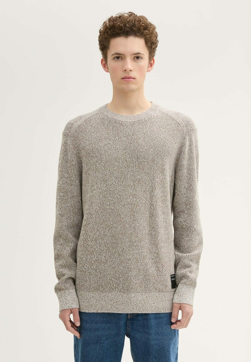 Hellbrauner Strickpullover mit Rundhalsausschnitt und gerippten Bündchen, der ein strukturiertes Muster und ein schwarzes Markenetikett am Saum aufweist.