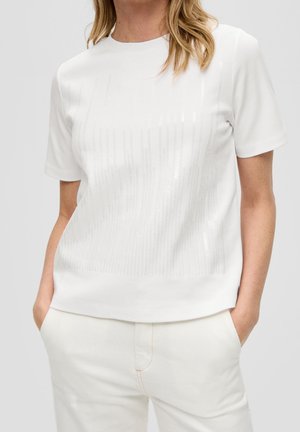 Frau trägt ein weißes kurzärmeliges Oberteil mit dezenten vertikalen Paillettenstreifen, kombiniert mit weißen Hosen, Hände in den Taschen.