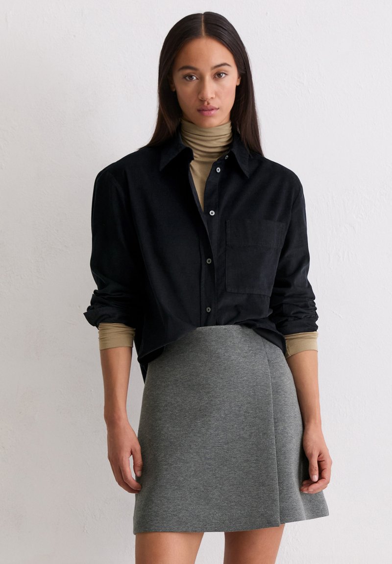 Chemise noire à boutons avec une poche poitrine, portée sur un col roulé beige et associée à une jupe grise ajustée. Textures lisses partout.
