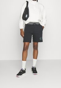 Black polyester shorts with cuffed hems and a round logo, paired with a white half-zip pullover and black sneakers, featuring green accents.
