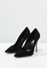 Ett par svarta mocka högklackade skor med spetsiga tår och slimma metalliska stilettklackar på en vit yta med enkel bakgrund.