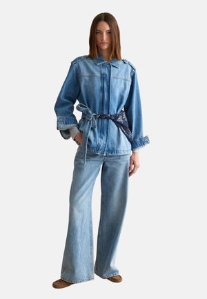 Vrouw die een lichtblauw spijkerjasje draagt met een blauw patroonriem, bijpassende wijde jeans en bruine mocassins, staand tegen een effen achtergrond.