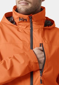 Jasně oranžová nepromokavá bunda s vysokým límcem, předním zipem a náprsní kapsou. Má texturovanou vnitřní podšívku a nastavitelné manžety.
