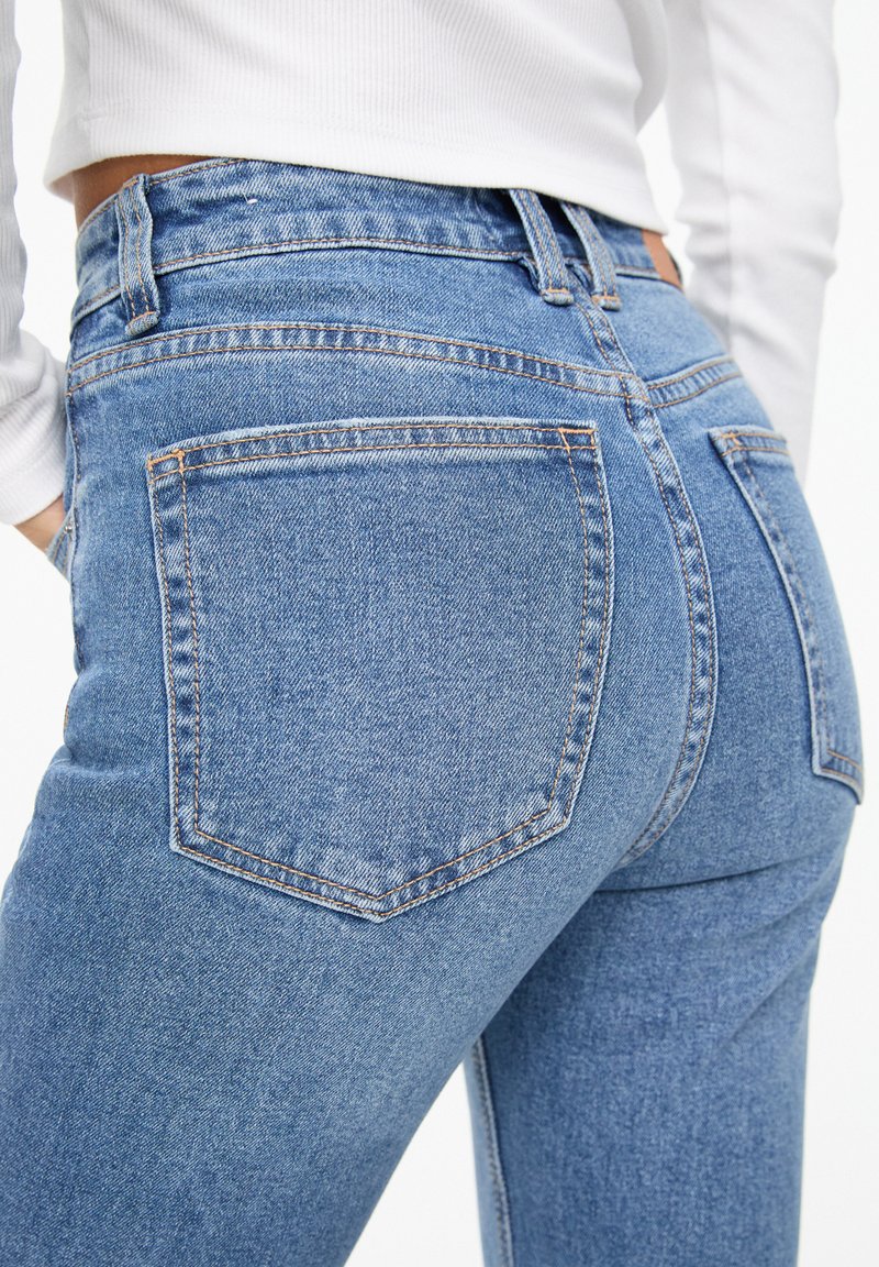 Jean en denim bleu au design ajusté, comprenant cinq poches, avec un léger délavage et des coutures orange.