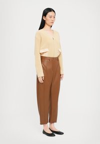 Maglione beige con zip, texture a coste e tasche frontali; abbinato a pantaloni in finta pelle marrone con vestibilità comoda e gamba dritta.