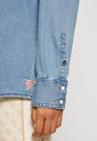 Manica di camicia in denim blu chiaro con due bottoni a pressione bianchi sul polsino e piccolo logo Guess rosso e bianco vicino al polso su tessuto beige a motivo.