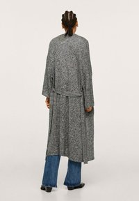 Longue robe grise tissée avec des manches larges, une ceinture à la taille et une surface texturée, portée sur un jean évasé bleu avec des chaussures noires.