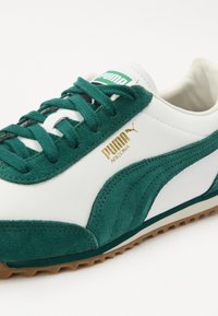 Grüne Wildleder- und weiße Leder-Sneaker mit einem strukturierten Design, ausgestattet mit goldenen Logo-Akzenten und grünen Schnürsenkeln auf einer Gummisohle.