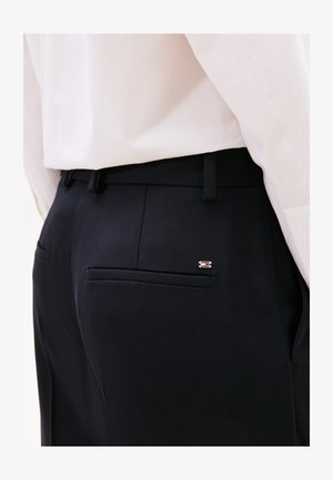 Pantaloni tailored blu con frontale piatto e tasche laterali, caratterizzati da un piccolo logo metallico accanto alla tasca posteriore. Tessuto liscio.