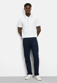 Weißes Polohemd mit einem kontrastierenden schwarzen Kragen, kombiniert mit blauen Hosen und weißen Sneakers. Glattes Material und taillierte Passform.