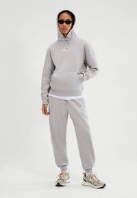 Grijze hoodie met kangoeroezak en trekkoord, gecombineerd met bijpassende joggingbroek. Voorzien van oranje logo. Model draagt een zonnebril en sneakers.