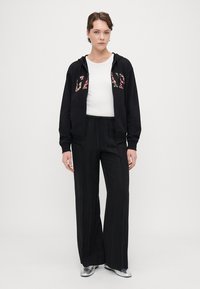 Črna zip-up kapuca z cvetličnim napisom "GAP", nošena nad belo majico, v kombinaciji s črnimi širokimi hlačami in metalno srebrnimi čevlji.