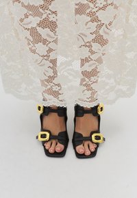 Schwarze Leder-Sandalen mit quadratischen Zehen, ausgestattet mit zwei großen goldenen Schnallen als Akzent. Getragen mit einem transparenten Spitzenoverlay.