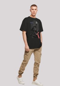 Svart bomull T-skjorte med et grafisk bilde av en karakter med et rødt lyssverd, kombinert med beige cargo-bukser og hvite sneakers.