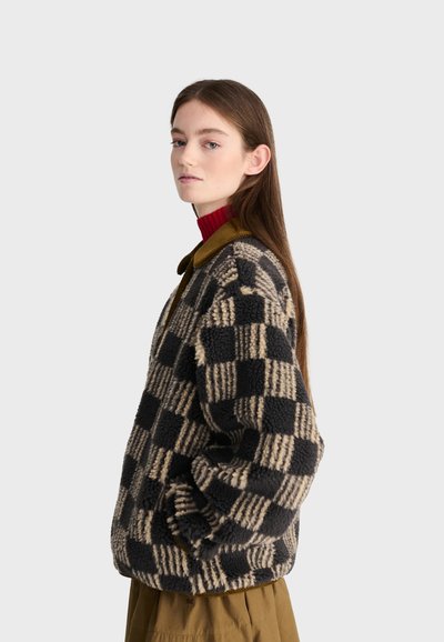 Jeune femme aux longs cheveux bruns portant une veste polaire à carreaux noire et beige et un pull à col roulé rouge, debout de profil.