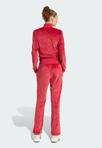 Crveni velur trenirku s prilagođenom jaknom i odgovarajućim hlačama. Tkanina ima mekanu, glatku teksturu, s rebrastim rubovima i pojasom.