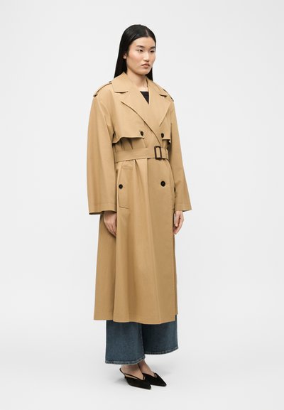 Γυναίκα στέκεται φορώντας ένα μακρύ μπεζ καμπαρντίνα με ζώνη πάνω από φαρδιά μπλε τζιν παντελόνια και μαύρες γόβες με αιχμηρή μύτη και λουρί πίσω, σε απλό λευκό φόντο.