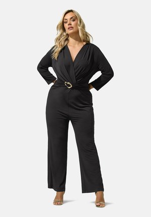 Femme aux longs cheveux blonds portant une combinaison-pantalon noire portefeuille avec une boucle de ceinture dorée et des talons ouverts beiges, debout, les mains sur les hanches.