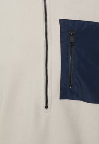 Nærbillede af beige fleecejakke med lodret sort lynlås og mørkeblå lynlås-lomme på brystet til højre.