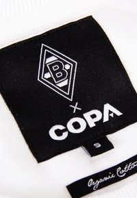 La etiqueta de tela negra presenta un logo blanco con una "B" y franjas. Acompañada del texto "x COPA". La etiqueta indica la talla "S" y el material "algodón orgánico."