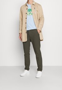 Chaqueta de camisa beige a la altura de la rodilla, camiseta gráfica azul con texto verde, pantalones olivo de corte ajustado y zapatillas blancas. Diseño minimalista, sin patrones.