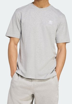 Mann trägt hellgraues Adidas-T-Shirt mit kurzen Ärmeln und passende Shorts, steht mit den Händen in den Taschen vor einem einfarbigen Hintergrund.