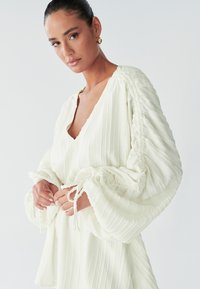 Een vrouw met een gladde huid draagt een getextureerde witte blouse met pofmouwen, terwijl ze met een zachte uitdrukking de koordjes om haar middel vasthoudt.
