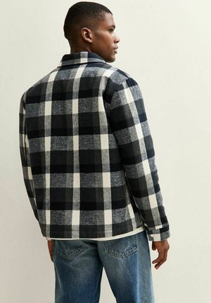Man in een zwart-wit-grijs geblokte shirtjack, staande in een zijaanzicht, gecombineerd met blauwe denim jeans tegen een effen achtergrond.
