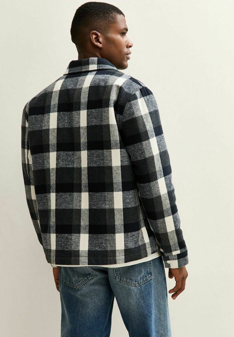 Man in een zwart-wit-grijs geblokte shirtjack, staande in een zijaanzicht, gecombineerd met blauwe denim jeans tegen een effen achtergrond.