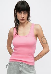 Camiseta de tirantes ajustada de color rosa con textura acanalada y tirantes anchos, con escote redondeado y detalle sutil del logo. Combinada con pantalones vaqueros gris claro.