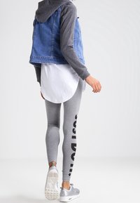 Denim vest with a grey long-sleeve shirt, white layered tunic, grey leggings featuring black text, and silver athletic shoes. Smooth textures.