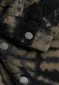 Giacca tie-dye nera e marrone con dettagli in metallo, tessuto testurizzato e cuciture in evidenza su colletto e polsini.