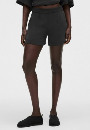 Kvinde iført sorte højlivs-shorts med forreste syninger, sorte slip-on sko, står mod en ensfarvet lys baggrund.