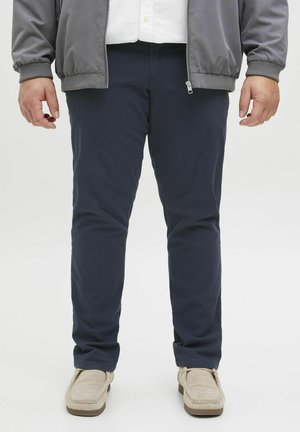 Marinefarbene Chinos mit glatter Textur, slim fit und Fronttaschen. Kombiniert mit beigen Slippern. Graue Bomberjacke teilweise sichtbar.