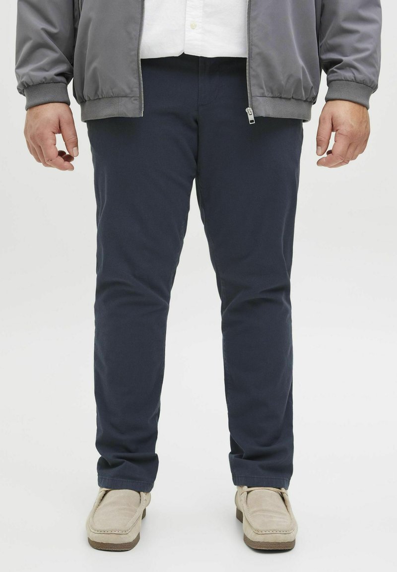 Marineblauwe chino's met een gladde textuur, slim fit en zijzakken. Gecombineerd met beige instapschoenen. Grijze bomberjack gedeeltelijk zichtbaar.