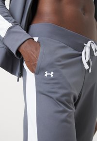Personne à la peau foncée portant un pantalon de survêtement gris Under Armour avec des bandes blanches sur les côtés, main dans la poche, et une manche de veste raccourcie visible.