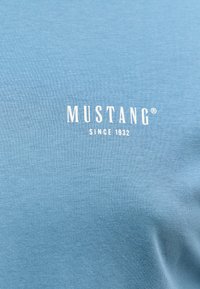 Jasnoniebieski bawełniany t-shirt z białym tekstem marki "MUSTANG" oraz "SINCE 1932". Miękka tekstura z gładkim wykończeniem. Bez wzorów ani grafik.