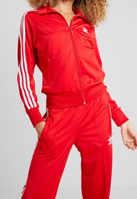 Röd träningsdräkt i glatt och glänsande tyg. Innehåller en zip-jacka med vita ränder på ärmarna och matchande byxor med sidofickor.