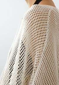 Ramie i górna część ramienia w lu�źno plecionej beżowej siateczkowej bluzie na czarnym podkoszulku, na jednolitym tle.