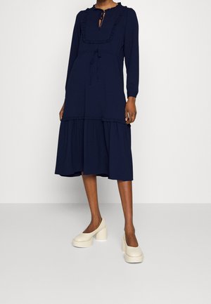 Vrouw draagt een marineblauwe midi-jurk met lange mouwen en ruches, gecombineerd met crèmekleurige plateauhakken, staand tegen een effen lichte achtergrond.