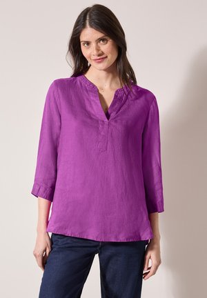 Camisa de lino morada con escote en V y mangas de tres cuartos, de corte relajado y con una textura sutil. Combinada con jeans oscuros.