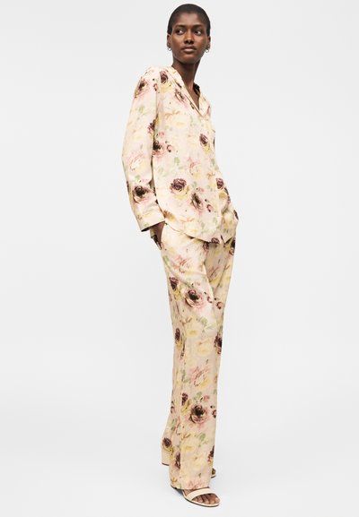 Naine kannab kreemikat lillemustriga pikade varrukatega särki ja sobivaid laiade säärtega pükse, seisab käed taskutes ühtlase tausta ees.