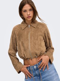 Jeune femme aux longs cheveux bruns portant une veste courte en daim marron avec fermeture éclair et un jean bleu, les mains reposant près de la ceinture.